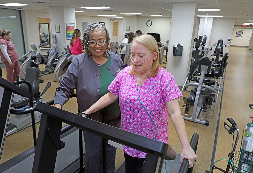 A smiling nurse helping a patient walk on a treadmill as part of their exercise therapy.