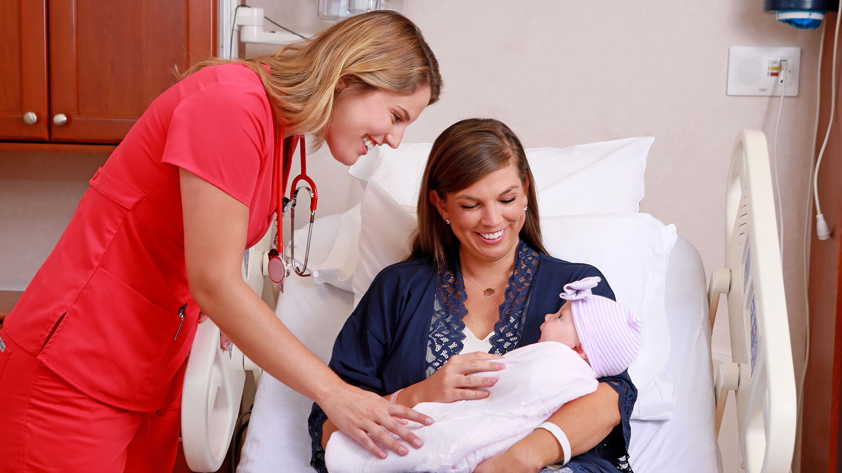 Nurse standing next to a mother sitting in a hospital bed holding a swaddled newborn.