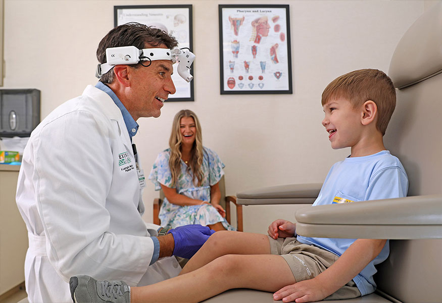 A smiling pediatric ear, nose and throat doctor speaking with a smiling child. A smiling mother watches in the background.